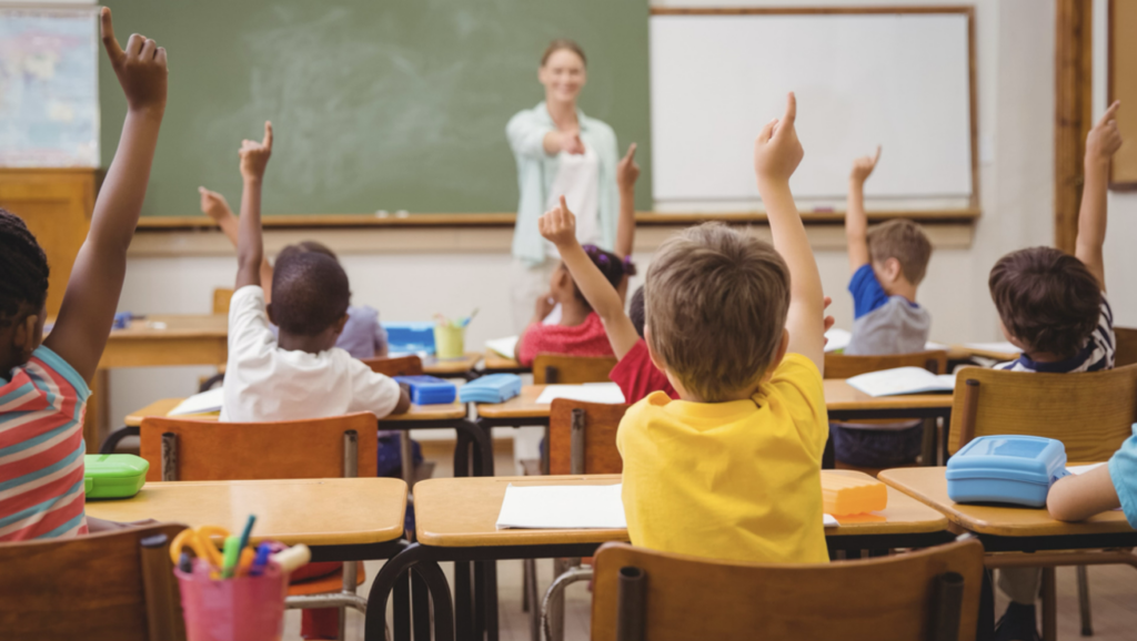 Skole elever rekker opp hånden i et klasserom, og en lærer peker på en av elevene. Foto.