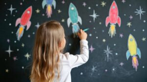 Liten jente tegner raketter og stjerner med kritt på tavlen. Jenta lager fargerik kunst inspirert av verdensrommet. Foto