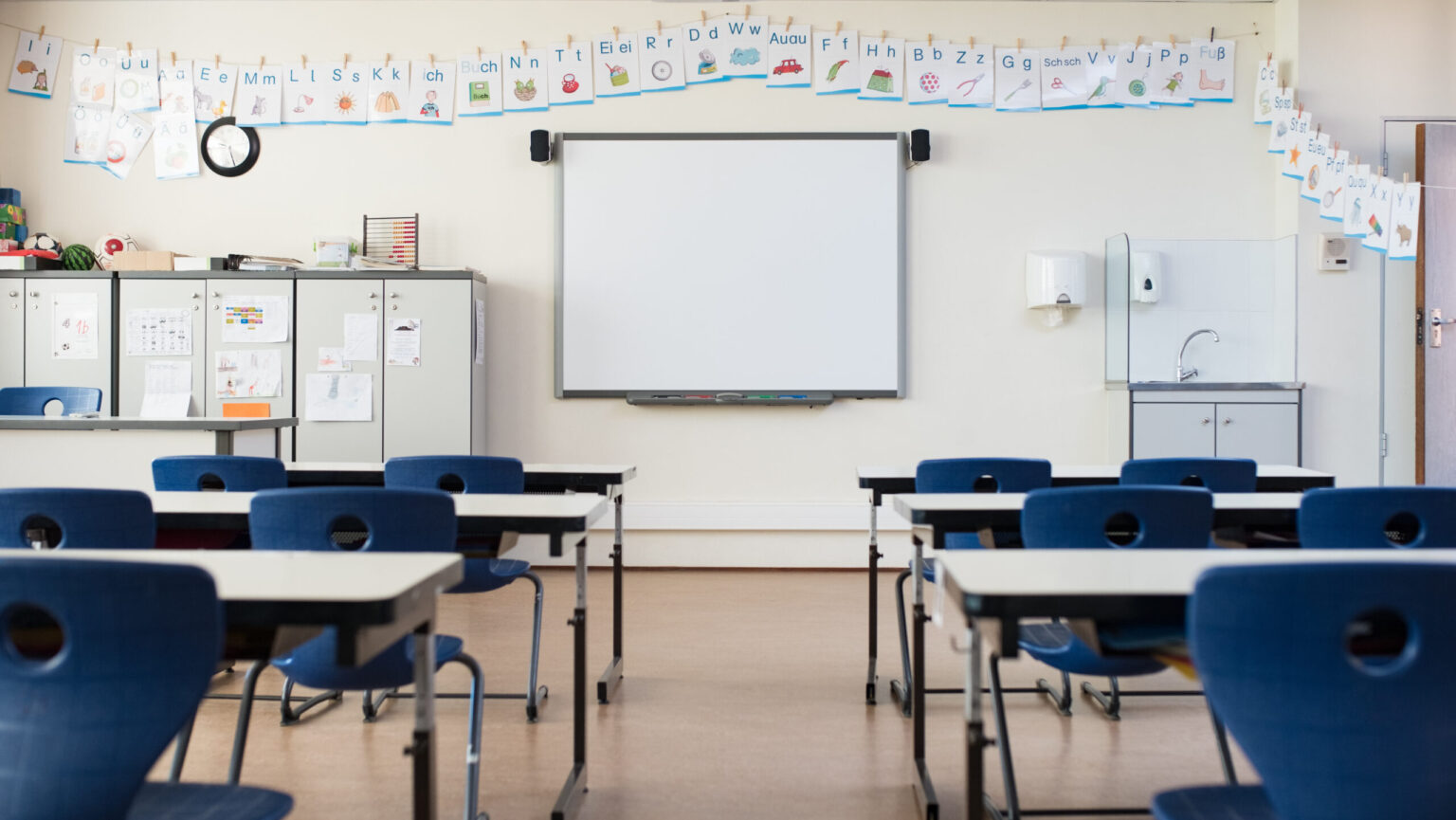 Skolepulter og stoler i et tomt, moderne klasserom. Tomt klasserom i barneskole med tavle og projektor. Klasserom for småskoletrinnet med smarttavle og alfabet på veggen. Foto.