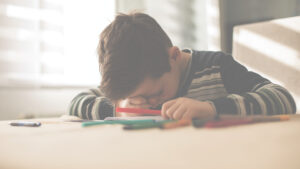 Et barn sitter ved et bord og tegner eller skriver med tusjer. Barnet lener hodet ned mot armen og konsentrerer seg om arket foran seg. Flere fargede tusjer ligger spredt på bordet, og mykt dagslys kommer inn gjennom et vindu i bakgrunnen. Foto. Jot something down