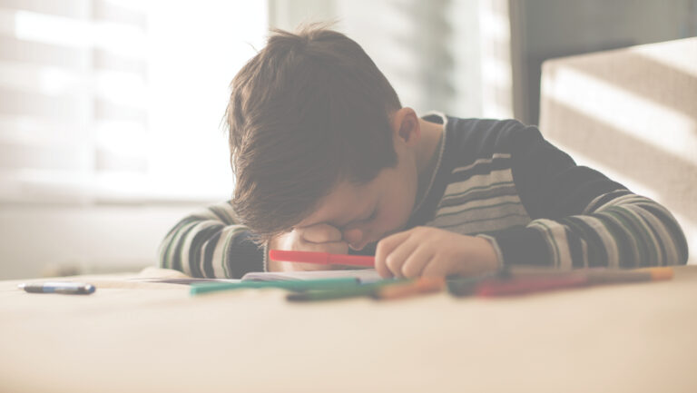 Et barn sitter ved et bord og tegner eller skriver med tusjer. Barnet lener hodet ned mot armen og konsentrerer seg om arket foran seg. Flere fargede tusjer ligger spredt på bordet, og mykt dagslys kommer inn gjennom et vindu i bakgrunnen. Foto. Jot something down