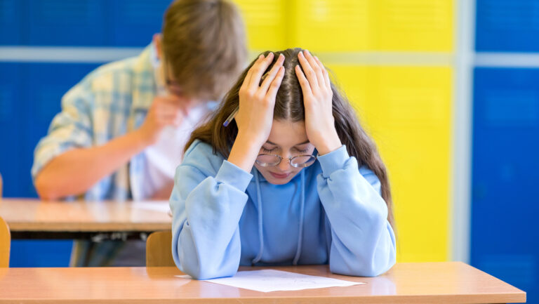 Bekymret kvinnelig elev sitter ved pulten i klasserommet og ser ned, med hodet i hendene. I bakgrunnen sitter en annen elev. Fotto