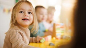 Små barn sitter rundt et bord i barnehagen. En liten jente i forgrunnen er i fokus og smiler. Foto.