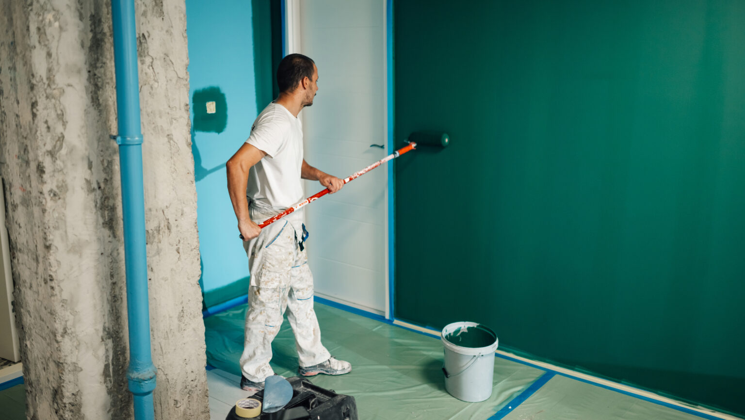 Profesjonell maler bruker en malerulle og påfører grønn maling på en vegg i et rom under oppussing av bolig. Foto.
