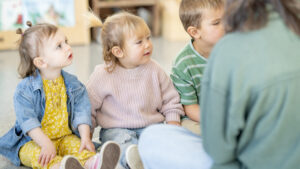 En gruppe barn sitter på klasseromsgulvet og lytter oppmerksomt til en voksen. Gruppen inkludert jente i gult, jente i rosa og en gutt. Foto