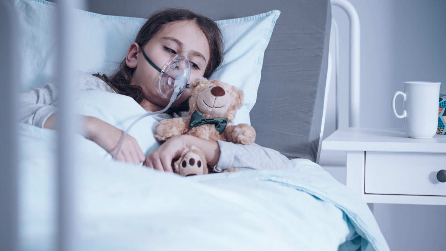 Barn med cystisk fibrose ligger i sykehusseng med oksygenmaske, mens det holder et kosedyr. Foto.