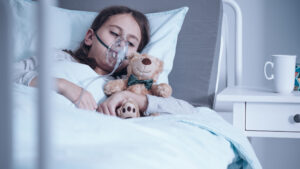 Barn med cystisk fibrose ligger i sykehusseng med oksygenmaske, mens det holder et kosedyr. Foto.